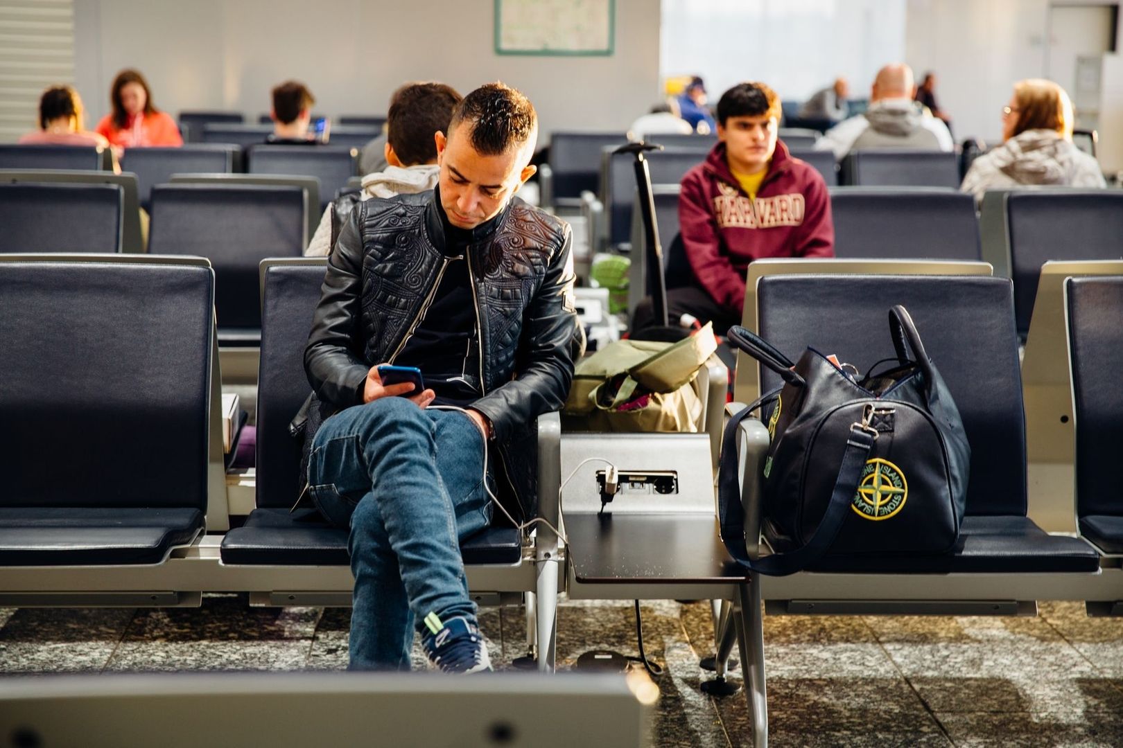 Passagiere warten mit Gepäck in einem Wartebereich und nutzen ein Smartphone